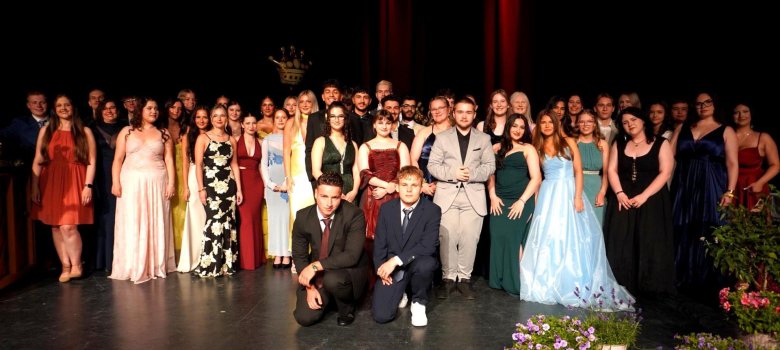 Gruppenfoto vom Abiball einer Schule: Etwa 45 festlich gekleidete Abiturient*innen stehen auf einer Bühne vor dunklem Hintergrund und rotem Bühnenlicht. Die jungen Erwachsenen tragen elegante Abendkleider, Anzüge und festliche Frisuren. Im Vordergrund knien zwei Schüler in Anzug und Krawatte. Die Stimmung wirkt feierlich und stolz. Der Bühnenrand ist mit Blumen geschmückt, im Hintergrund ist eine goldene Krone als Dekoration zu sehen.