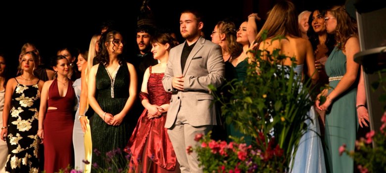Abitur 2025 Eine festlich gekleidete Gruppe junger Erwachsener steht auf einer Bühne und blickt zur Seite, als würde sie auf die nächste Programmansage warten. Die Personen tragen elegante Kleider und Anzüge, im Vordergrund sind große Blumentöpfe mit bunten Blumen zu sehen. Die Bühne ist dunkel ausgeleuchtet, die Gruppe wird von warmem Scheinwerferlicht angestrahlt, was eine feierliche Atmosphäre erzeugt.