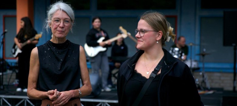 Frau Knowles (stellvertretende Schulleiterin) und Clara Henschke (Schülersprecherin) stehen nebeneinander und lächeln in die Kamera. Im Hintergrund spielt eine Schüler*innen-Band auf einer kleinen Bühne.