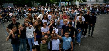 Eine große Gruppe Schüler*innen steht auf einem Schulhof dicht beieinander, einige halten Urkunden oder Pokale in den Händen. Im Vordergrund zeigen mehrere Kinder einen Pokal in die Kamera. Im Hintergrund sitzt eine große Menge Publikum auf Stühlen.