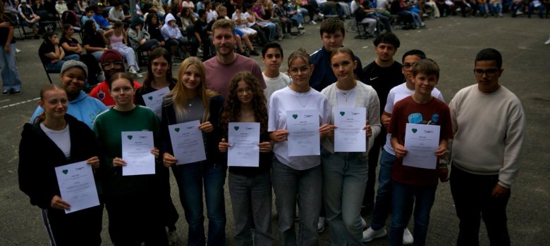 Schüler*innen der Gesamtschule Gronau stehen in zwei Reihen auf dem Schulhof. Sie halten Zertifikate in den Händen und lächeln in die Kamera. In ihrer Mitte steht Herr Nöthe, der sie als verantwortlicher Lehrer begleitet. Im Hintergrund sitzt das Publikum auf dem Schulhof.