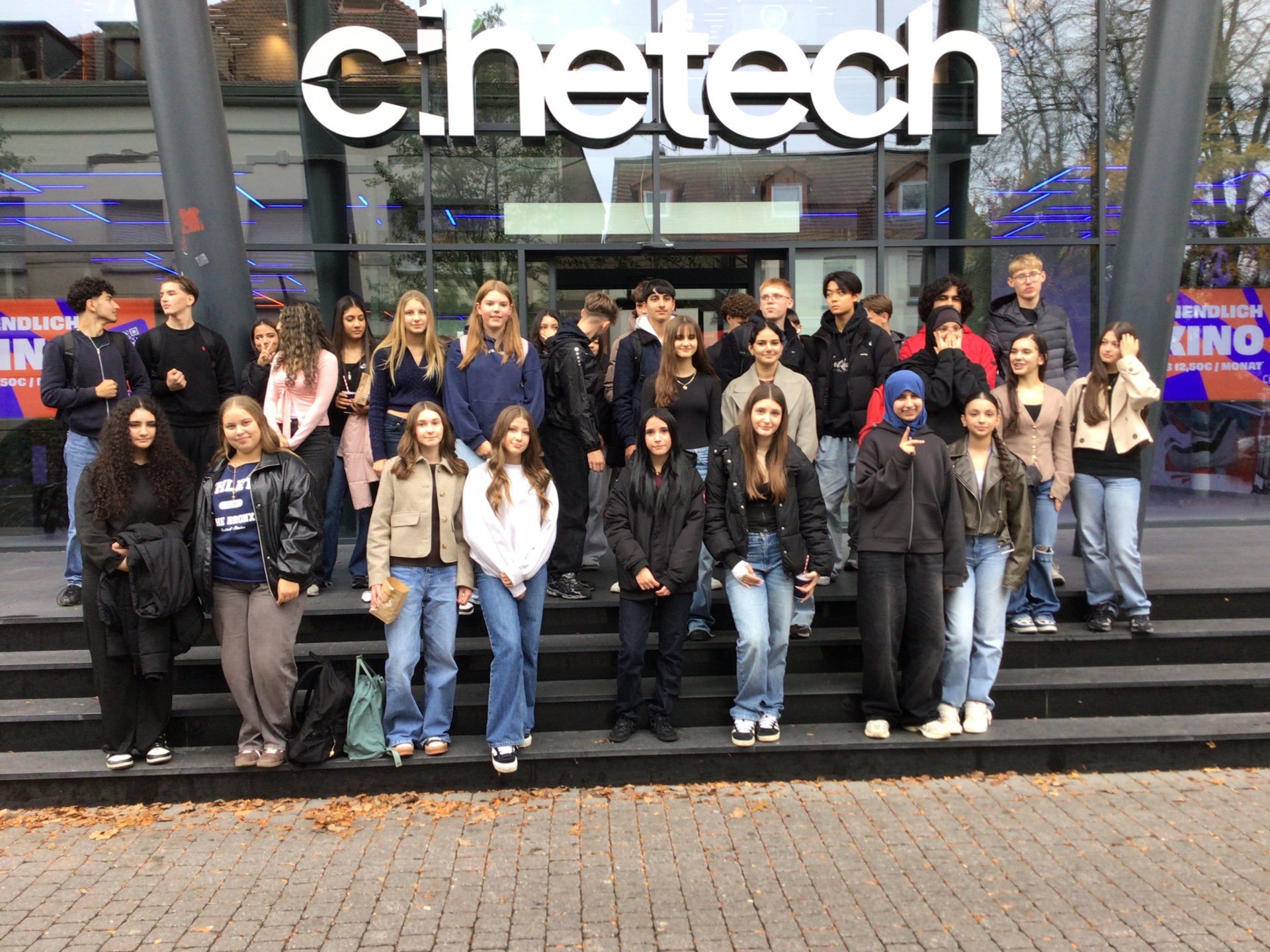 Eine große Gruppe von Schüler*innen der Gesamtschule Gronau und des Stedelijk Lyceum Enschede Zuid steht gemeinsam auf den Treppen vor dem Cinetech-Kino in Gronau. Die Jugendlichen posieren für ein Gruppenfoto nach einem gemeinsamen Austausch zum Thema Nachhaltigkeit. Im Hintergrund ist die Glasfassade des Kinos mit dem leuchtenden Schriftzug „Cinetech“ zu sehen.