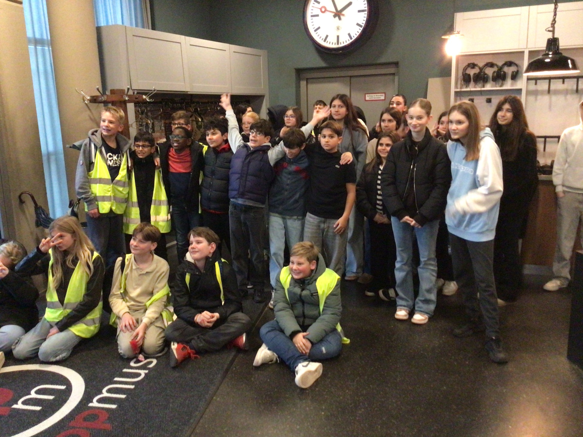Auf dem Foto ist eine Gruppe von rund 25 Schülerinnen in einem Eingangsbereich zu sehen. Einige sitzen vorne auf dem Boden, andere stehen dahinter dicht beieinander. Mehrere Schülerinnen tragen gelbe Warnwesten, die anderen Jacken oder Hoodies. Viele schauen in Richtung Kamera, einige lächeln oder winken. Hinter der Gruppe hängt eine große, runde Uhr an der Wand, darunter ist eine geschlossene Tür. Links im Bild sieht man eine Garderobe mit vielen Jacken und ein paar Regenschirmen, rechts Regale mit Kopfhörern und eine Hängelampe. Der Raum wirkt wie das Foyer eines Museums oder einer Ausstellung.