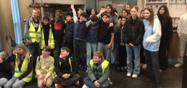 Auf dem Foto ist eine Gruppe von rund 25 Schülerinnen in einem Eingangsbereich zu sehen. Einige sitzen vorne auf dem Boden, andere stehen dahinter dicht beieinander. Mehrere Schülerinnen tragen gelbe Warnwesten, die anderen Jacken oder Hoodies. Viele schauen in Richtung Kamera, einige lächeln oder winken. Hinter der Gruppe hängt eine große, runde Uhr an der Wand, darunter ist eine geschlossene Tür. Links im Bild sieht man eine Garderobe mit vielen Jacken und ein paar Regenschirmen, rechts Regale mit Kopfhörern und eine Hängelampe. Der Raum wirkt wie das Foyer eines Museums oder einer Ausstellung.