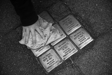 Schwarz-Weiß-Foto in Nahaufnahme: Eine Schüler*innen-Hand der Gesamtschule Gronau wischt am 4. November 2025 mit einem Tuch über mehrere Stolpersteine, sodass die Namen und Daten der jüdischen Opfer im Rahmen der Woche der Erinnerung wieder klar zu erkennen sind.