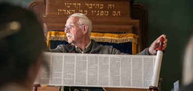 Ein älterer Ehrenamtlicher mit Kippa sitzt vor dem Toraschrein der Synagoge in Enschede und hält eine geöffnete Nachbildung einer Torarolle mit mehreren Spalten hebräischen Textes in den Händen, während davor Schüler*innen der Gesamtschule Gronau sitzen.