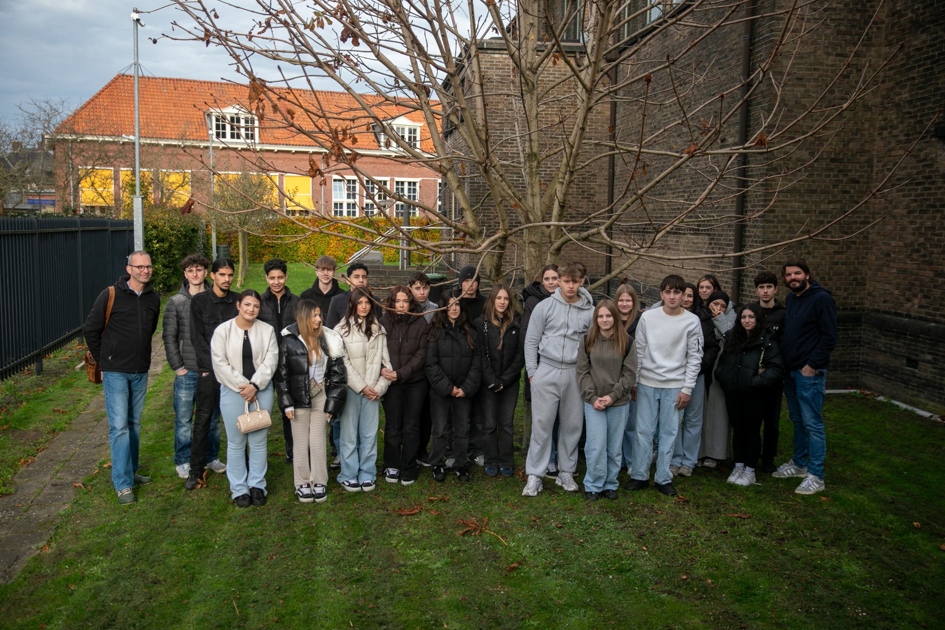Eine große Gruppe von Schüler*innen des 9./10. Jahrgangs der Gesamtschule Gronau steht gemeinsam mit zwei Lehrkräften auf der Rasenfläche neben der Synagoge in Enschede; hinter ihnen sind ein kahler Baum, die Backsteinmauer der Synagoge und Wohnhäuser mit roten Dächern zu sehen.