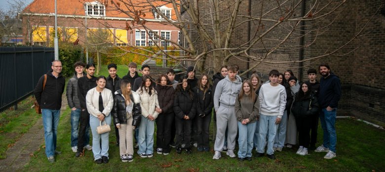 Eine große Gruppe von Schüler*innen des 9./10. Jahrgangs der Gesamtschule Gronau steht gemeinsam mit zwei Lehrkräften auf der Rasenfläche neben der Synagoge in Enschede; hinter ihnen sind ein kahler Baum, die Backsteinmauer der Synagoge und Wohnhäuser mit roten Dächern zu sehen.