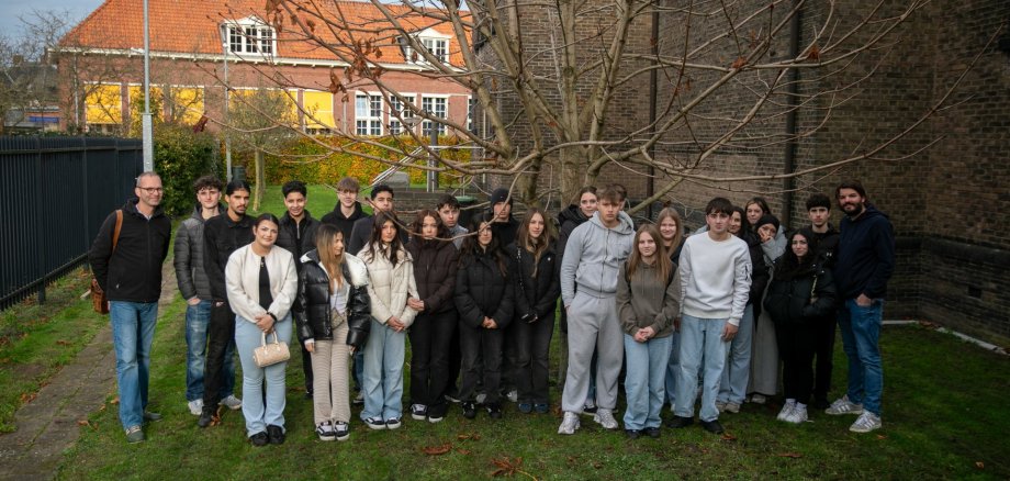 Eine große Gruppe von Schüler*innen des 9./10. Jahrgangs der Gesamtschule Gronau steht gemeinsam mit zwei Lehrkräften auf der Rasenfläche neben der Synagoge in Enschede; hinter ihnen sind ein kahler Baum, die Backsteinmauer der Synagoge und Wohnhäuser mit roten Dächern zu sehen.