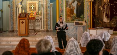 In der reich bemalten syrisch-orthodoxen Kirche in Gronau steht die Schülerin Hala mit weißem Spitzenschleier vor dem Altar und spricht zu den auf Kirchbänken sitzenden Schüler*innen des 9./10. Jahrgangs, die teilweise ebenfalls Schleier tragen; im Hintergrund sind Kreuz, Ikonen und Kerzen zu sehen.