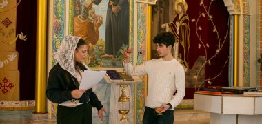Vor einer großen Heiligenikone in der syrisch-orthodoxen Kirche in Gronau steht die Schülerin Hala mit Spitzenschleier und einem Blatt Papier neben einem Mitschüler, der ein goldenes Weihrauchfass an langen Ketten hochhält und in der anderen Hand ein kleines Gefäß trägt; gemeinsam zeigen sie der Schüler*innengruppe liturgische Gegenstände des Gottesdienstes.