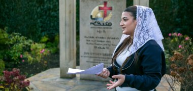 Im Garten der syrisch-orthodoxen Kirche in Gronau steht die Schülerin Hala mit weißem Spitzenschleier und einem Blatt Papier in der Hand und spricht; hinter ihr befindet sich das steinerne Mahnmal „Sayfo Syriac Genocide“, das mit Kreuzsymbol und mehrsprachiger Inschrift an den Genozid an rund 500.000 Aramäer*innen im Jahr 1915 erinnert.