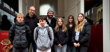 Eine kleine Gruppe von fünf Schüler*innen der 9./10. Klasse und zwei Lehrkräften der Gesamtschule Gronau steht in Winterkleidung in einem Hauseingang in der Innenstadt, blickt in die Kamera und nimmt im Rahmen der Woche der Erinnerung 2025 am Projekt „Stolpersteine putzen“ teil.