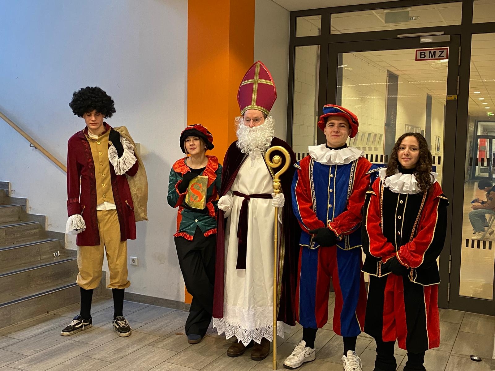 Eine als Sinterklaas verkleidete Person mit Mitra und Bischofsstab steht mit vier als traditionelle Begleiterinnen verkleideten Schüler*innen im Flur der Gesamtschule Gronau. Die Gruppe trägt farbenfrohe Kostüme und bereitet den Besuch der Klassen des fünften Jahrgangs vor.