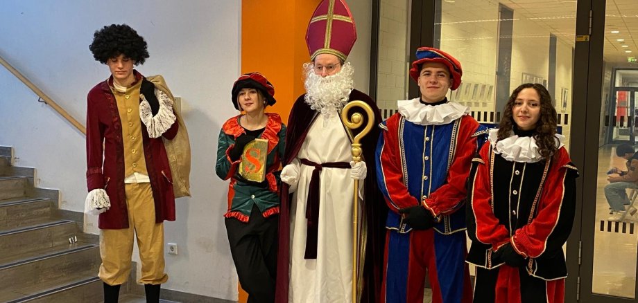 Eine als Sinterklaas verkleidete Person mit Mitra und Bischofsstab steht mit vier als traditionelle Begleiterinnen verkleideten Schüler*innen im Flur der Gesamtschule Gronau. Die Gruppe trägt farbenfrohe Kostüme und bereitet den Besuch der Klassen des fünften Jahrgangs vor.