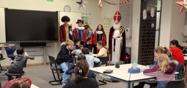 In einem weihnachtlich dekorierten Klassenraum sitzen Schüler*innen des fünften Jahrgangs an Gruppentischen, während Sinterklaas und seine bunt kostümierten Helfer*innen vorne im Raum stehen. Die Kinder schauen neugierig nach vorn und freuen sich über den Besuch und die kleinen Geschenke.