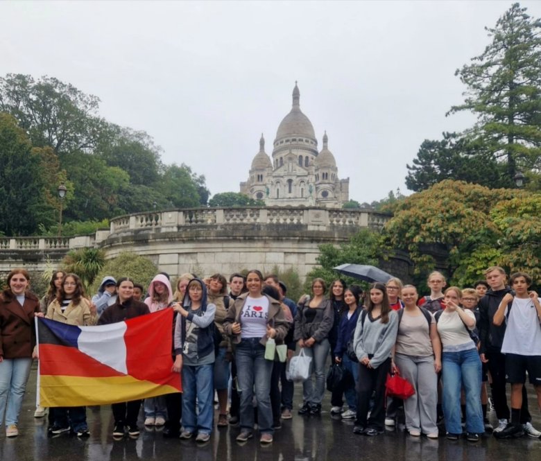 Schülergruppe bei einer Frankreichfahrt vor der Basilika Sacré-Cœur auf dem Montmartre in Paris. Einige Jugendliche halten eine deutsch-französische Flagge. Im Hintergrund sind die helle Kuppelkirche und die begrünte Terrasse zu sehen.