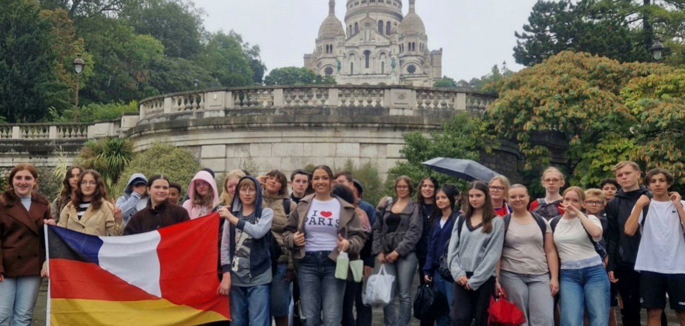 Schülergruppe bei einer Frankreichfahrt vor der Basilika Sacré-Cœur auf dem Montmartre in Paris. Einige Jugendliche halten eine deutsch-französische Flagge. Im Hintergrund sind die helle Kuppelkirche und die begrünte Terrasse zu sehen.