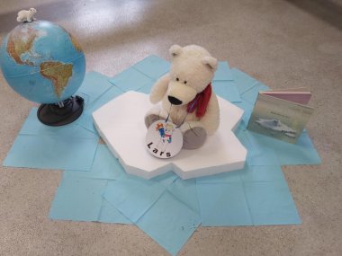 Klassentier Eisbär sitzt auf einer Scholle, neben ihm sind ein Globus und ein Buch zu sehen