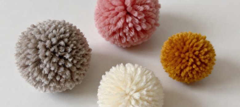 Das Bild zeigt 4 Pom-Poms in grau, rosa, weiß und orange auf einem weißen Tisch.
