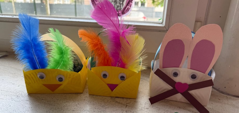 Das Foto zeigt drei selbstgebastelte Osterkörbchen. Einen weiß/rosanen Hasen und zwei gelbe Küken mit bunten Federn.